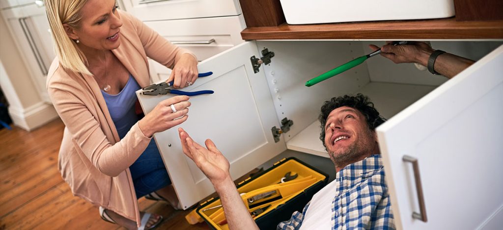 man under sink shutting off water valves with woman handing him pliers
